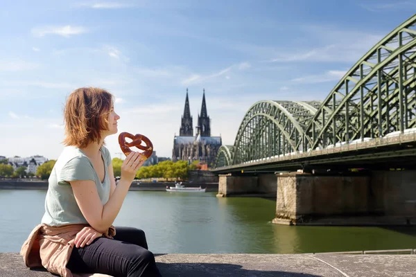 Young woman eats traditional pretzel sitting on embankment of Rhine on background of Cologne Cathedral and Hohenzollern Bridge in Koel, Germany — Stock Photo, Image