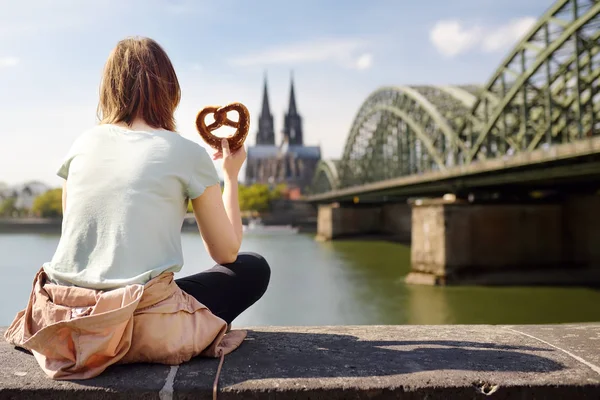 Tourist eats traditional pretzel sitting on embankment of Rhine on background of Cologne Cathedral and Hohenzollern Bridge in Koel, Germany — Stock Photo, Image