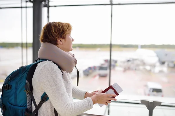 Mulher com uma mochila e um travesseiro de viagem olha no avião na janela na área de espera do aeroporto . — Fotografia de Stock