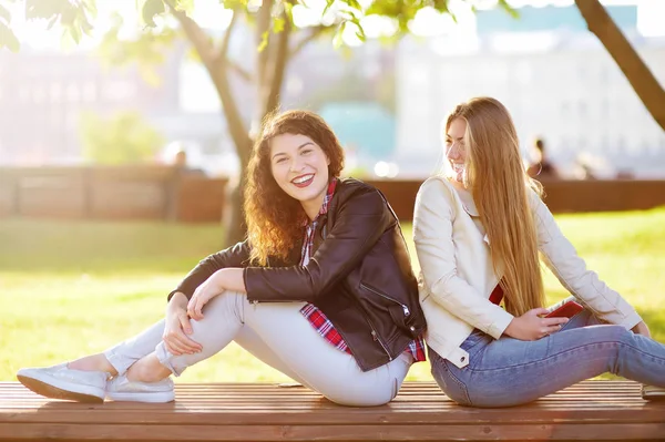 Zwei schöne junge Frauen unterhalten sich auf einer Bank im sonnigen Park. — Stockfoto