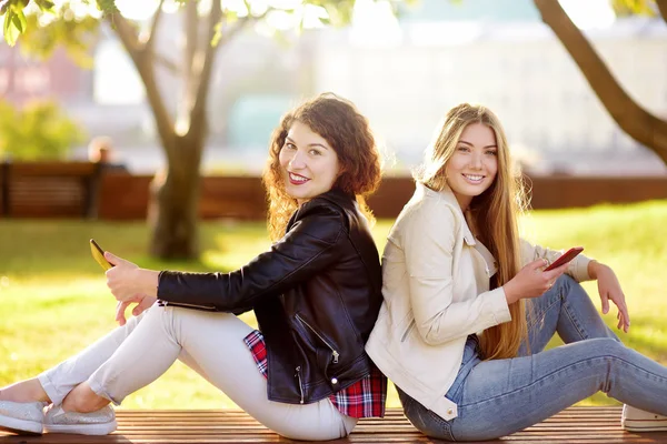 Duas belas mulheres jovens conversando enquanto sentadas em um banco no parque ensolarado . — Fotografia de Stock