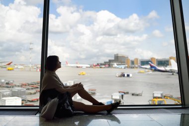 Bir sırt çantası ve bir seyahat yastığı ile kadın oturan ve havaalanının bekleme alanında pencerede uçağa bakıyor.