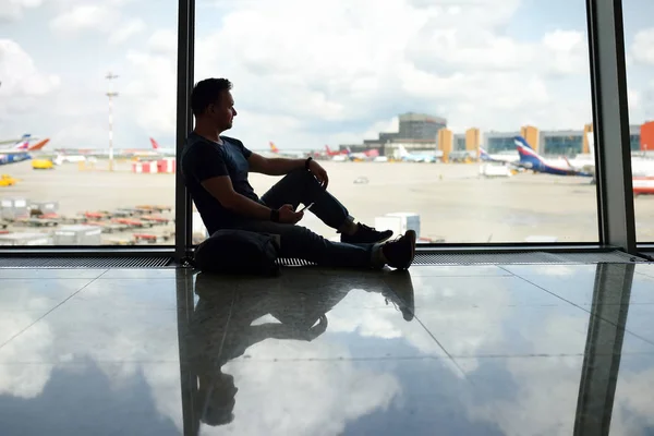 Mannen met een rugzak zitten en kijken op het vliegtuig in venster op de wachtruimte van de luchthaven. — Stockfoto