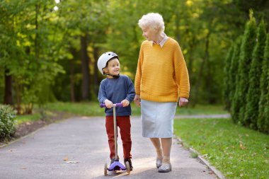 Beautiful granny and her little grandchild walking together in autumn park. Boy riding by scooter.