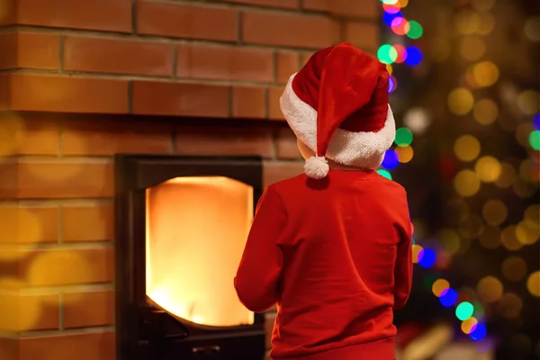 Little child dressed in a Santa hat and festive costume looks at fireplace on Christmas eve at home — Stock Photo, Image