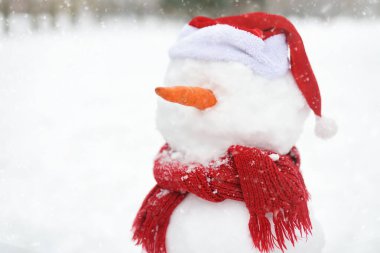 El yapımı eşarplı bir kardan adamın yakın plan fotoğrafı, Noel Baba şapkası ve karlı bir parkta havuç burnu..
