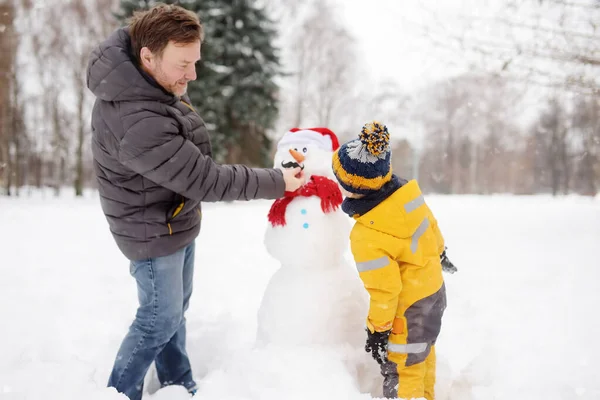 Menino com o pai a construir boneco de neve no parque nevado. Atividades ao ar livre lazer com crianças no inverno . — Fotografia de Stock