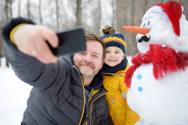 Malý chlapec a jeho otec berou selfie na pozadí sněhuláka ve zasněženém parku. Aktivní volný čas venku s dětmi v zimě. — Stock fotografie
