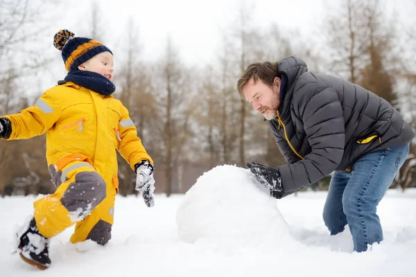Malý chlapec se svým otcem staví sněhuláka ve sněžném parku. Aktivní volný čas venku s dětmi v zimě. — Stock fotografie