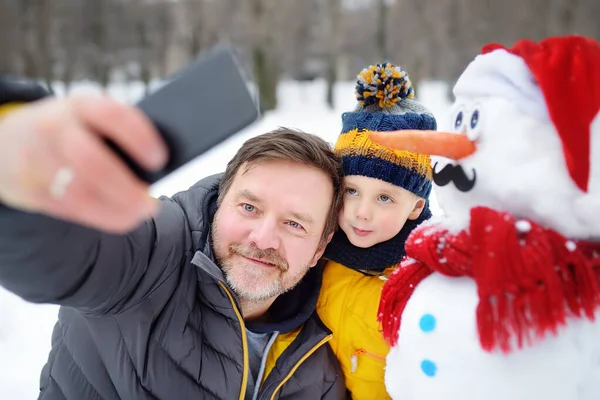 Mały chłopiec i jego ojciec robią sobie selfie na tle bałwana w zaśnieżonym parku. Aktywny wypoczynek na świeżym powietrzu z dziećmi w zimie. — Zdjęcie stockowe
