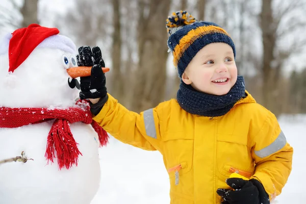 Chlapec buduje legračního sněhuláka. ? hild připevňuje mrkvový nos na sněhuláka ve zasněženém parku. — Stock fotografie