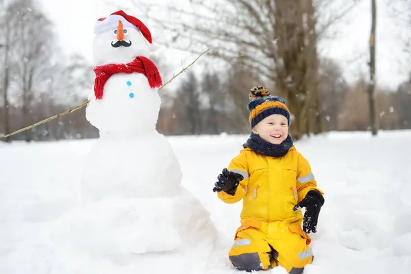 Malý chlapec si hraje s vtipným sněhulákem. Aktivní volný čas venku s dětmi v zimě. — Stock fotografie