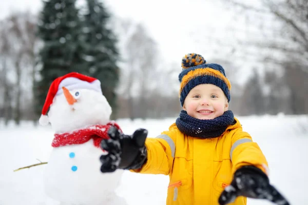 A kisfiú vicces hóemberrel játszik. Aktív szabadtéri kikapcsolódás gyerekekkel télen. — Stock Fotó