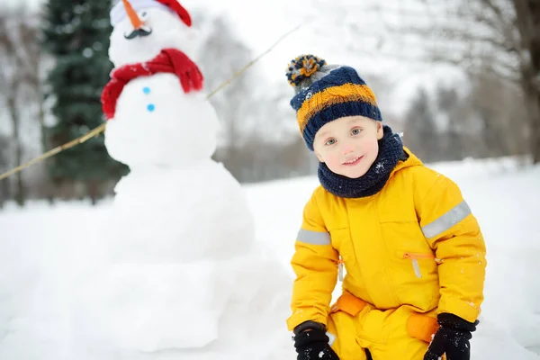 Malý chlapec si hraje s vtipným sněhulákem. Aktivní volný čas venku s dětmi v zimě. — Stock fotografie