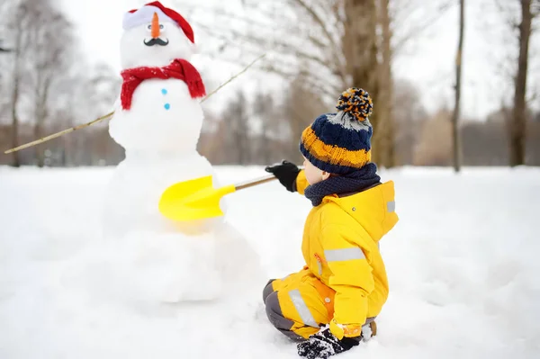 Mały chłopiec budujący bałwana z szalikiem i marchewkowym nosem w śnieżnym parku. — Zdjęcie stockowe