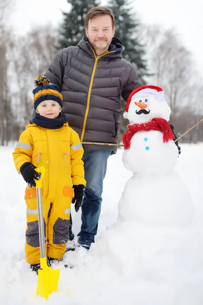 Niño pequeño con su padre construyendo muñeco de nieve en el parque nevado. Activo ocio al aire libre con niños en invierno . —  Fotos de Stock