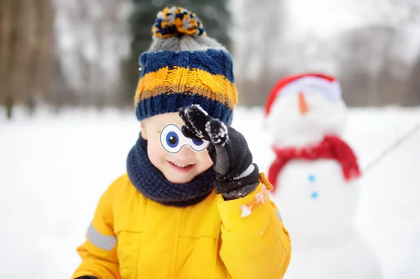 Mały chłopiec bawiący się zabawnym bałwanem. Jego oczy przymierza bałwan. Aktywny wypoczynek na świeżym powietrzu z dziećmi w zimie. — Zdjęcie stockowe