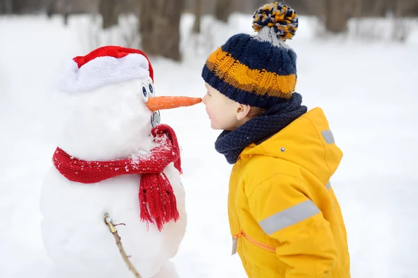 Little boy playing with funny snowman. Child reaches for a snowman's carrot nose and wants to bite. Active outdoors leisure with children in winter. — Stock Photo, Image