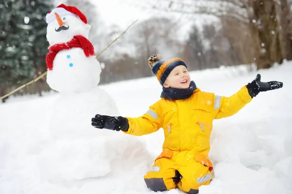 A kisfiú vicces hóemberrel játszik. Aktív szabadtéri kikapcsolódás gyerekekkel télen. — Stock Fotó