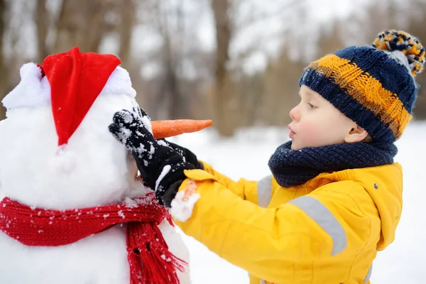 En liten pojke som bygger en rolig snögubbe. ♪ bygga fäster morot näsa till snögubbe i snöig park. — Stockfoto