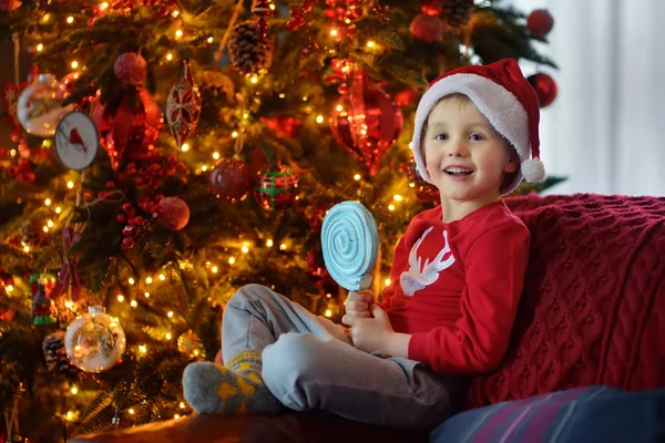 可爱的小男孩坐在圣诞树旁的沙发上,手里拿着大大的甜糖果. 圣诞假期快乐孩子的画像. — 图库照片