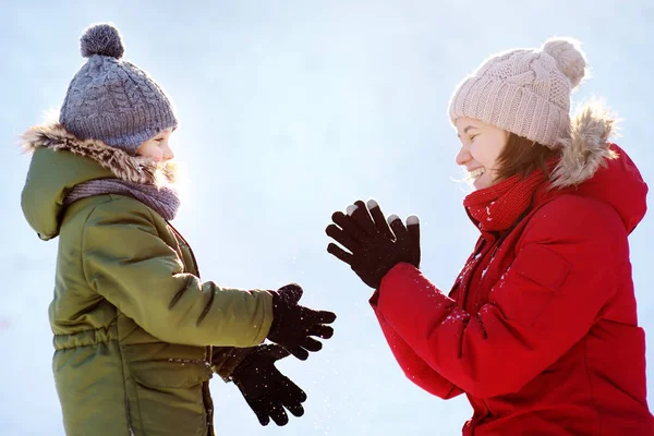 Menino bonito e jovem mãe jogando bolas de neve juntos durante o passeio no parque de inverno nevado. Diversão de inverno em família ao ar livre . — Fotografia de Stock