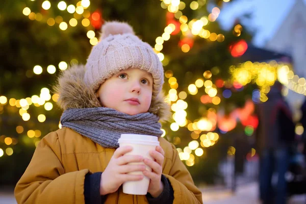 A kisfiú forró csokit vagy forralt bort iszik a nagy karácsonyfa közelében.. — Stock Fotó