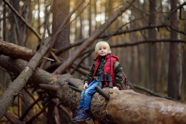 Маленький хлопчик-розвідник з біноклями під час походів в осінньому лісі. Дитина сидить на великому падаючому дереві і дивиться через бінокль . — стокове фото