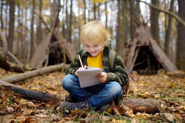 小男孩侦察兵正在森林里探路. 小孩坐在倒下的树上，在笔记本上写字。 孩子的后面是小茅屋. — 图库照片