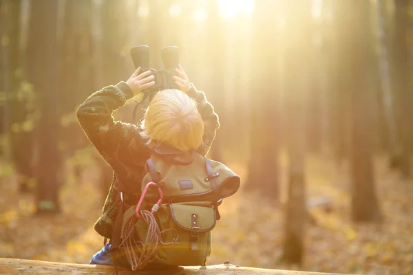在秋天的森林里徒步旅行时,小男孩用双筒望远镜侦察. 小孩坐在倒下的大树上，透过双筒望远镜望着. — 图库照片