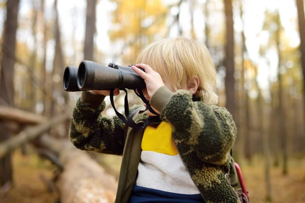 Malý skaut s dalekohledem během túry v podzimním lese. Dítě sedí na velkém padlém stromě a dívá se dalekohledem. — Stock fotografie