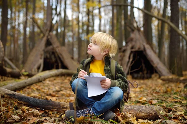 Malý skaut se orientuje v lese. Dítě sedí na padlém stromě a píše do notepadu. Teepee bouda na pozadí. — Stock fotografie