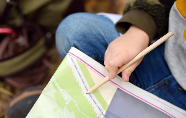 Il boy scout si sta orientando nella foresta. Bambino è seduto su un albero caduto e guardando sulla mappa sullo sfondo della capanna teepee . — Foto Stock