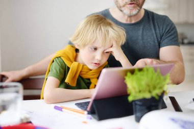 Baba çocuğuna evde ödev yapması için yardım ediyor. Yorgun çocuk bunu anlamıyor. Evde eğitim, uzaktan öğrenme, çevrimiçi eğitim, karantina sırasında çocuklar için uzaktan eğitim birçok ebeveyn için sorundur.