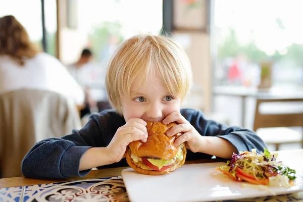 可愛いブロンドの男の子がファーストフード店で大きなハンバーガーを食べています 子供のための不健康な食事 ジャンクフード 太りすぎ問題の子 — ストック写真