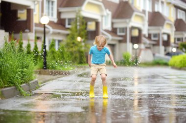 Küçük çocuk, yağmurlu bir yaz gününde küçük bir kasabada su birikintisine atlayan sarı lastik çizmeler giyiyor. Çocuk eğleniyor. Yağmurda çocuklar için açık hava oyunları.