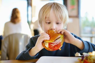 Fast food restoranında büyük hamburger yiyen sevimli sarışın çocuk. Çocuklar için sağlıksız bir yemek. Abur cubur. Aşırı kilolu çocuk.