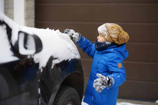 彼の父の車の上に横に新鮮な雪で遊んでかわいい子供 喜んでいる少年は雪のドリフトをノックダウン 子供は雪の嵐の後に車を掃除する親を助けます — ストック写真