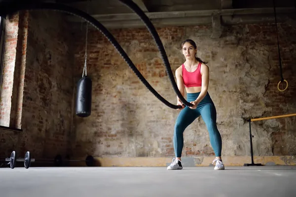 Young woman exercising with battle ropes at the gym. Strong female athlete doing crossfit workout with battle rope. Regular sports boosts immune system and promote good health and resistance to diseases. Healthy lifestyle