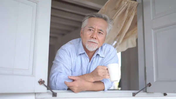 Senior asian man looking by window, holding cup of tea at home