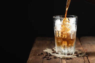 Buzlu kahve üzerinden uzun bir cam ve kahve çekirdekleri bir eski rustik ahşap tablo. Kopya alanı ile karanlık bir arka plan üzerinde soğuk yaz içeceği. Bir bardak içine bir kahve kabı üzerinden dökülen içki süreci.