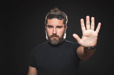 Siyah bir gömlek ve siyah bir arka plan Studio güneş gözlüğü sakallı ciddi adam. Kamera önünde duran ve stop sinyali sol onun palm ile gösterilen kendine güvenen adam. Metniniz için kopya alanı.