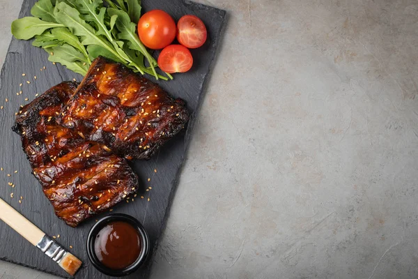 Primo piano di costolette di maiale alla griglia con salsa barbecue e rucola. Spuntino saporito a birra su un asse di pietra per limatura su sfondo concreto vecchio. Vista dall'alto con spazio di copia — Foto Stock