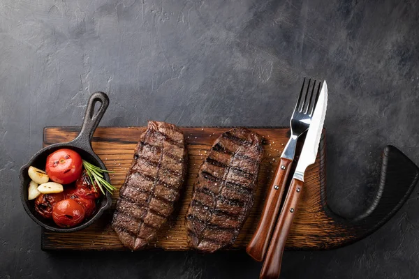 Closeup biftek üst bıçak sığır ırkları siyah yemeye hazır Angus ile ızgara domates, sarımsak ve ahşap bir gemide. Bitmiş çanak taş karanlık bir arka plan akşam yemeği için. Kopya alanı ile üstten görünüm.