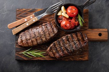 Closeup ready to eat steak Top Blade beef breeds of black Angus with grill tomato, garlic and on a wooden Board. The finished dish for dinner on a dark stone background. Top view clipart