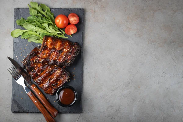 Primer plano de costillas de cerdo a la parrilla con salsa barbacoa y rúcula. La colación sabrosa a la cerveza sobre la piedra La tabla para la presentación sobre el fondo viejo de hormigón. Vista superior con espacio de copia — Foto de Stock