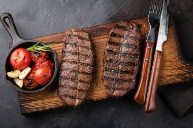Closeup biftek üst bıçak sığır ırkları siyah yemeye hazır Angus ile ızgara domates, sarımsak ve ahşap bir gemide. Bitmiş çanak taş karanlık bir arka plan akşam yemeği için. Üstten görünüm.