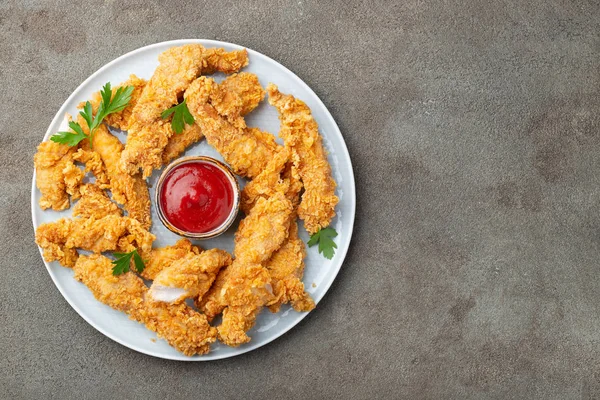 Breaded chicken strips with tomato ketchup on a white plate. Fast food on dark brown background. Top view with copy space.