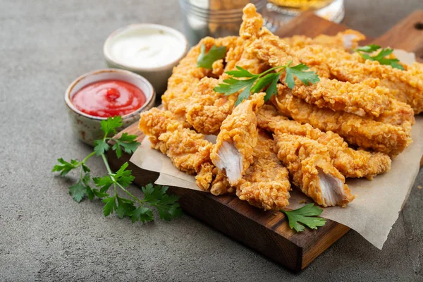 Breaded chicken strips with two kinds of sauces on a wooden Board. Fast food on dark brown background.