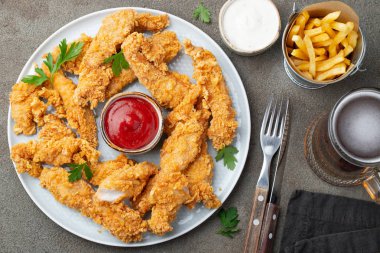 Breaded chicken strips with two kinds of sauces, fried potatoes and beer on a wooden Board. Fast food on dark brown background. Top view. clipart